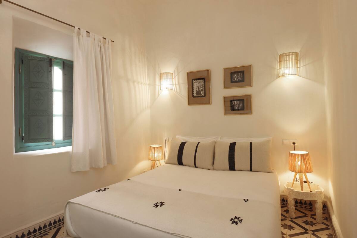 A bedroom with a bed, two bedside lamps, a window with curtains, and wall-mounted picture frames.