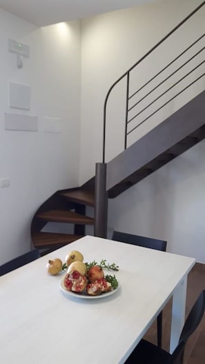 A dining area with a white table and a plate of food. A staircase with a metal railing is visible in the background.