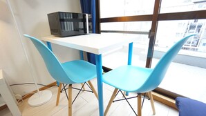 A room with a white table and two blue chairs, a microwave on the table, and a view through a large window. - Terry's Apartment Shinsaibashi I M04B (Osaka)
