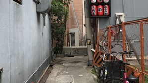 Ein schmaler Gang mit einem Fahrrad an der rechten Seite, einem roten Baustellenkegel und einem Gebäude mit einer Schautafel, die Speisen anbietet.