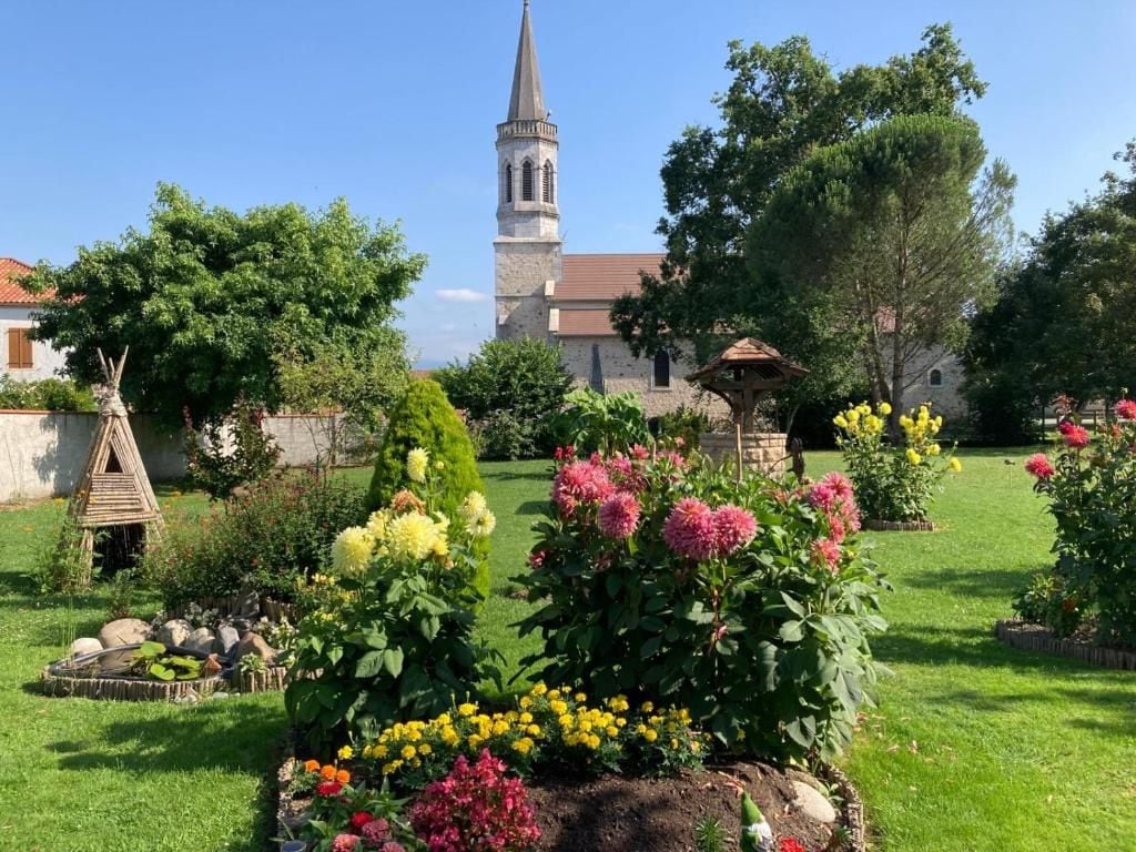 Un jardín con diversas flores y un pabellón, una iglesia con una aguja alta, y un cielo azul despejado.