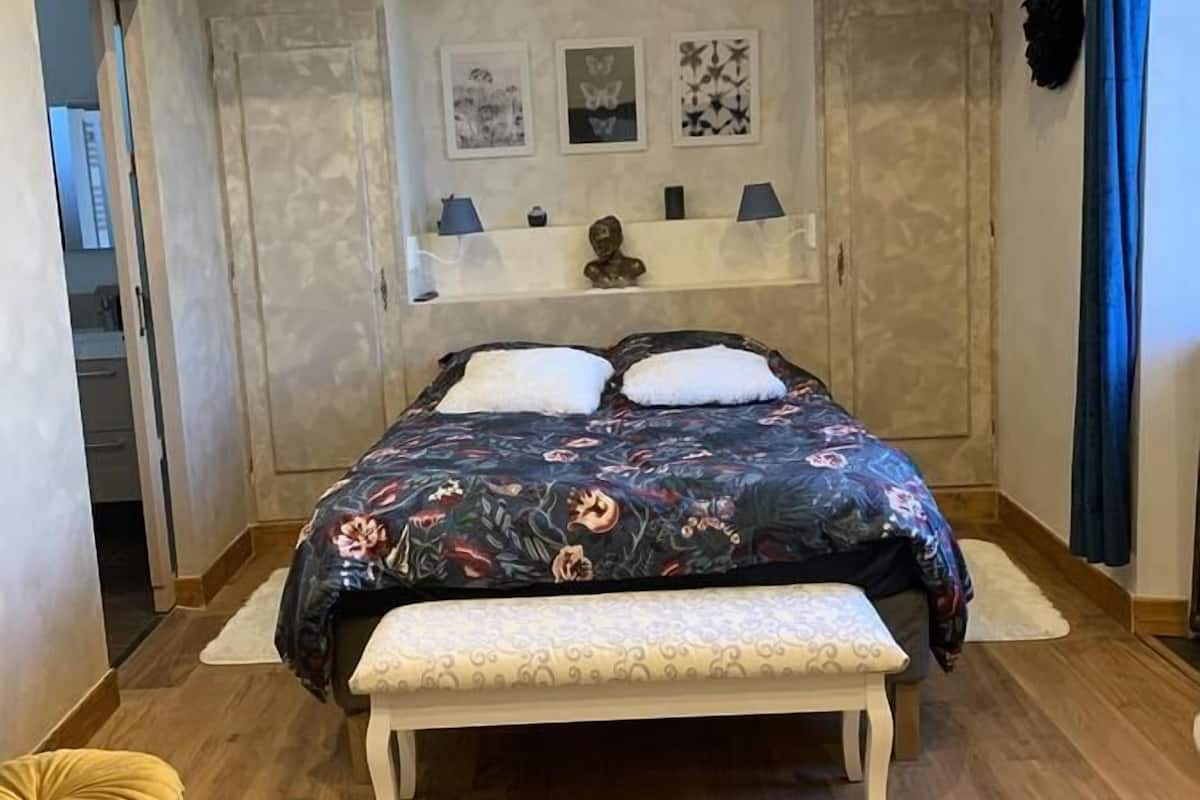 A bedroom with a large bed, a bench, and a small table. There are three wall-mounted shelves with decorative items.