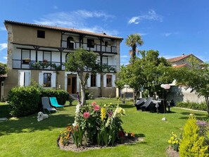 Un edificio de dos pisos con un jardín delantero, que incluye césped, flores y asientos al aire libre.