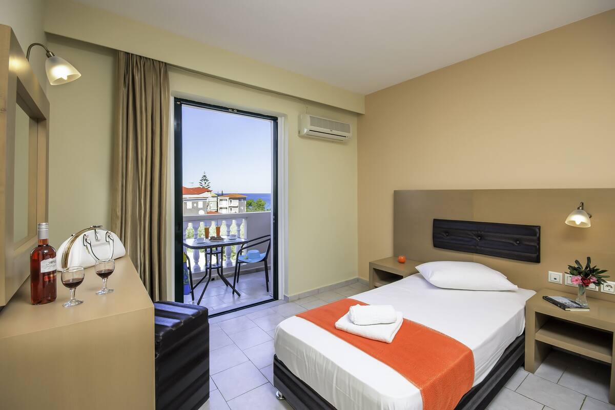 A hotel room with a bed, a desk with a bottle and glasses, a nightstand, and a view of a balcony with a table and chairs.