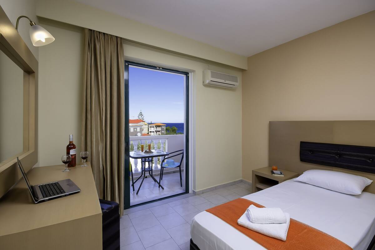 A hotel room with a bed, a desk with a laptop, a table with chairs, and a balcony with a view.