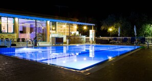 Zona de piscina nocturna con bar y área de estar, junto a un edificio con grandes ventanales.