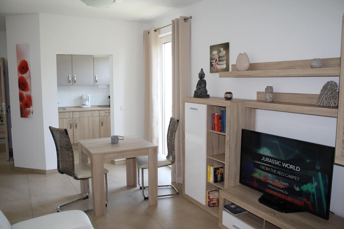 A modern living room with a flat-screen TV, wooden shelves, and a dining area with a table and chairs.