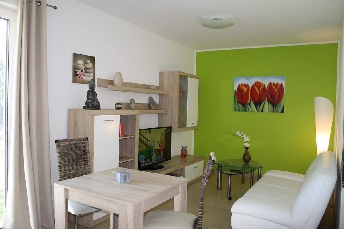 A modern living room with a wooden dining table, a white sofa, a TV, and a painting of tulips on the green wall.