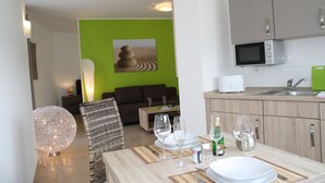 Una cocina moderna con microondas, pileta y muebles de color verde. Hay un comedor con una mesa preparada para dos personas, incluyendo copas de vino y platos.