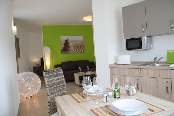 A modern kitchen with a microwave, sink, and green cabinets. There is a dining area with a table set for two, including wine glasses and plates. - Cloud 7 I (Heinsberg)