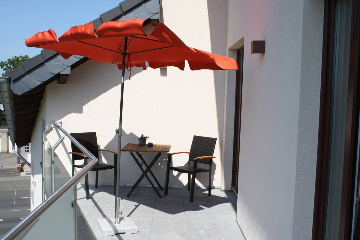 A balcony with a table and two chairs, an orange umbrella, and a potted plant.