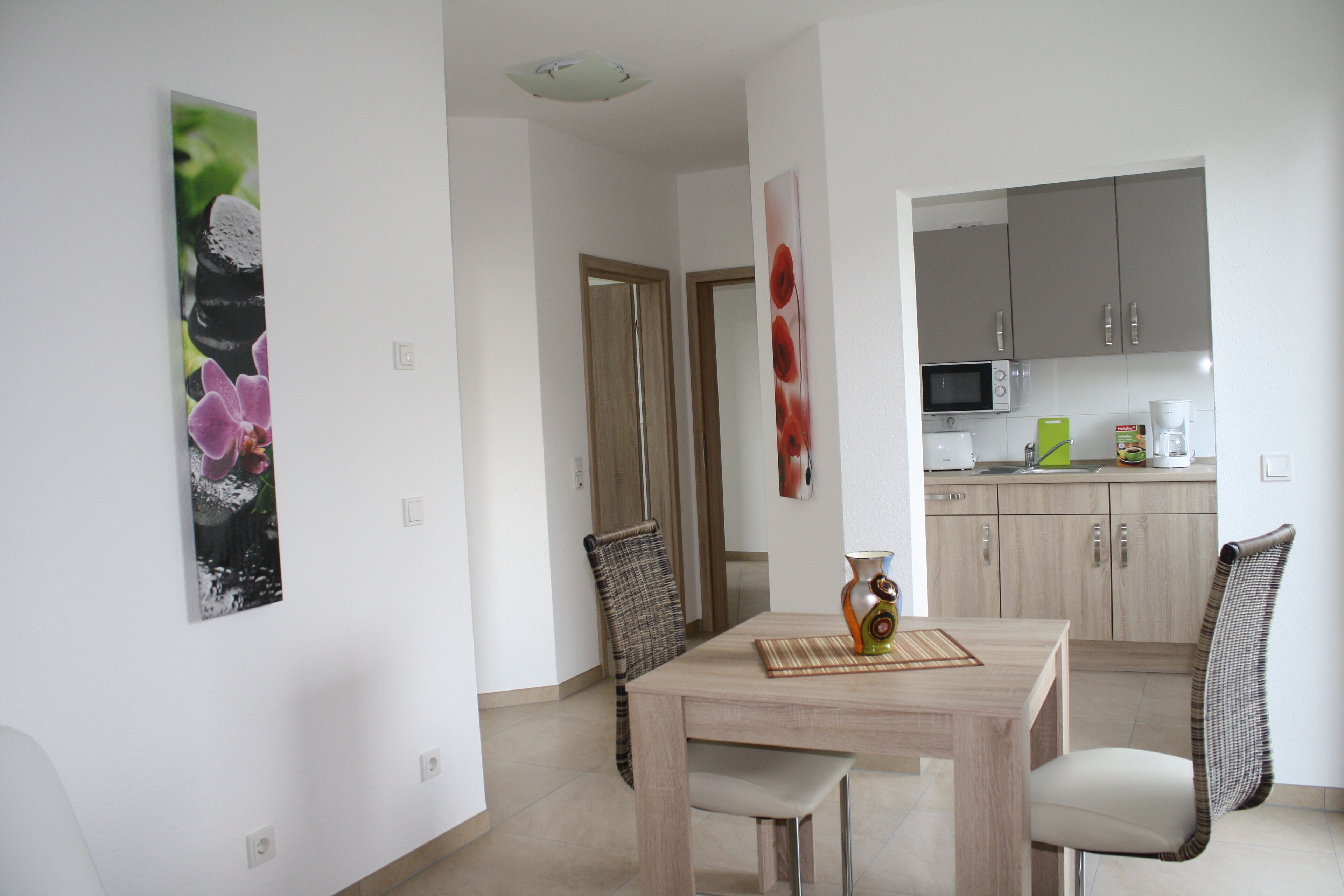 A dining area with a wooden table, two chairs, and a vase. There is a kitchen in the background with cabinets and appliances.