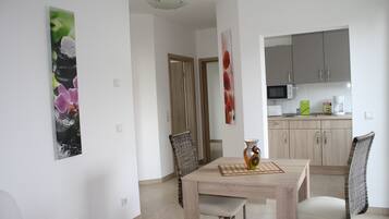 A dining area with a wooden table, two chairs, and a vase. There is a kitchen in the background with cabinets and appliances.