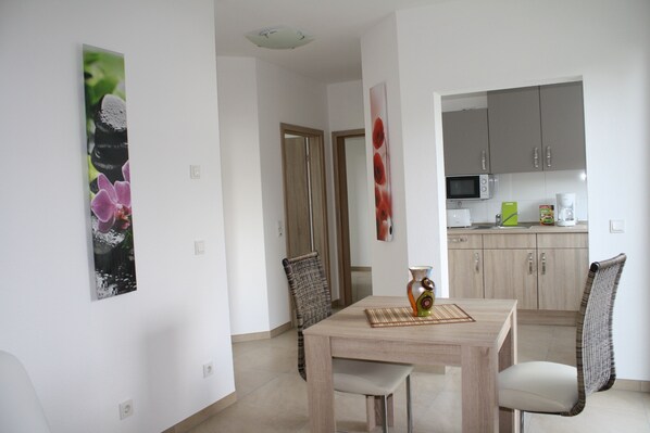 Un comedor con una mesa de madera, dos sillas y un florero. Se observa una cocina al fondo con armarios y electrodomésticos.