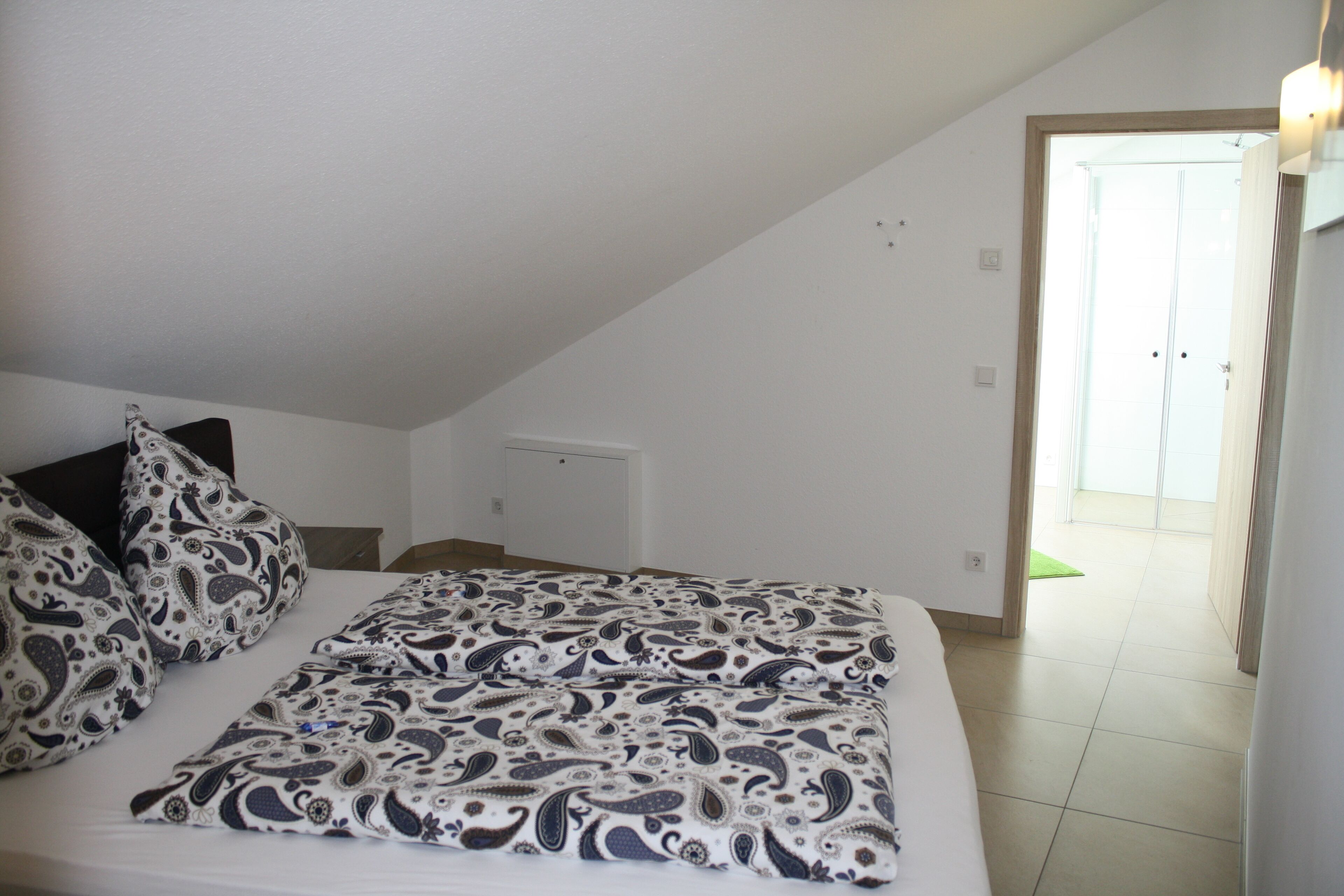 A bedroom with a bed, patterned pillows, a visible door, and a light switch on the wall.
