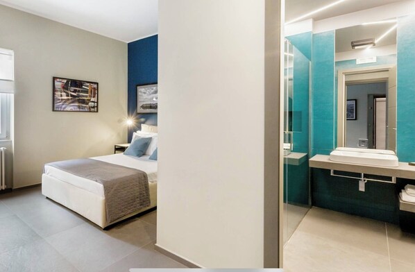 Un dormitorio con cama, mesita de noche y lámpara de noche. La habitación tiene una ventana, un cuadro enmarcado en la pared y una fuente de luz sobre la cama.