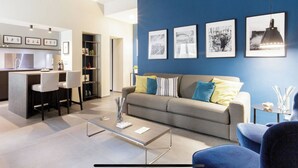 A modern living room with a sofa, coffee table, and chairs. There is a kitchen area with a counter and chairs. The room has blue walls and framed pictures on the wall. - Bluechiaia apartment- close to everything. (Napoli)