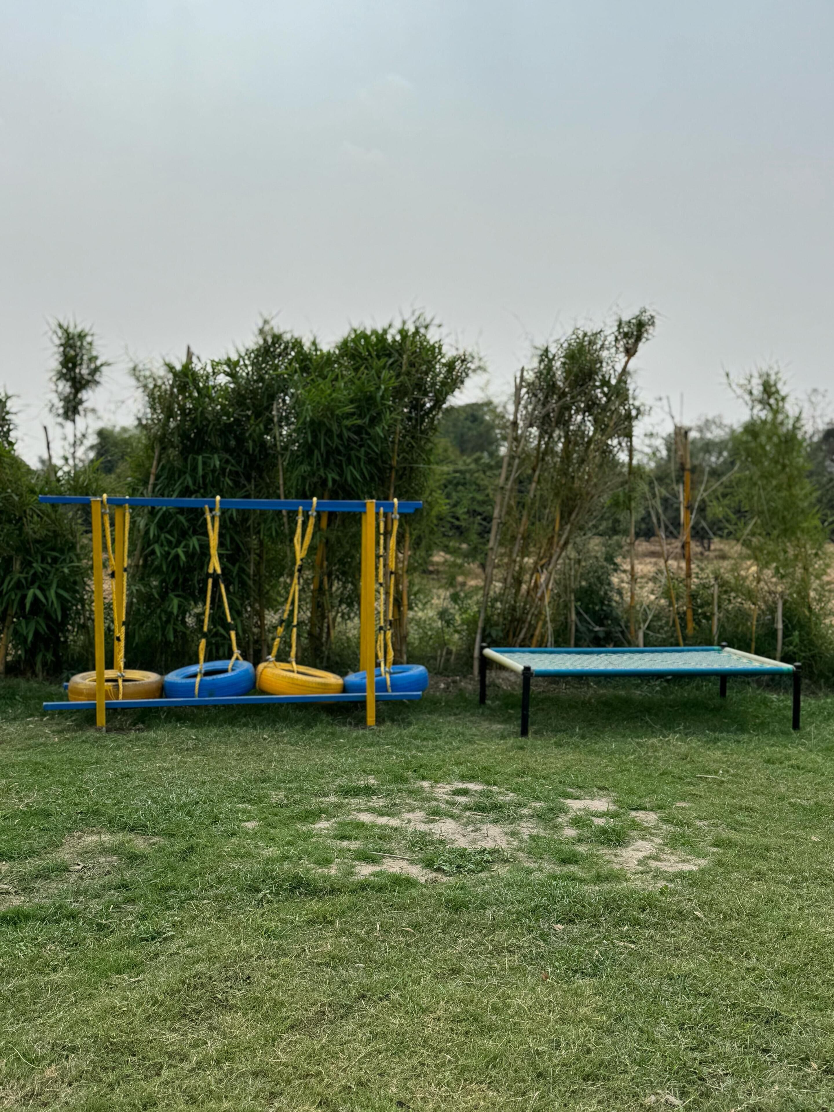 Un parque infantil con columpios y un trampolín en un área verde con árboles al fondo.