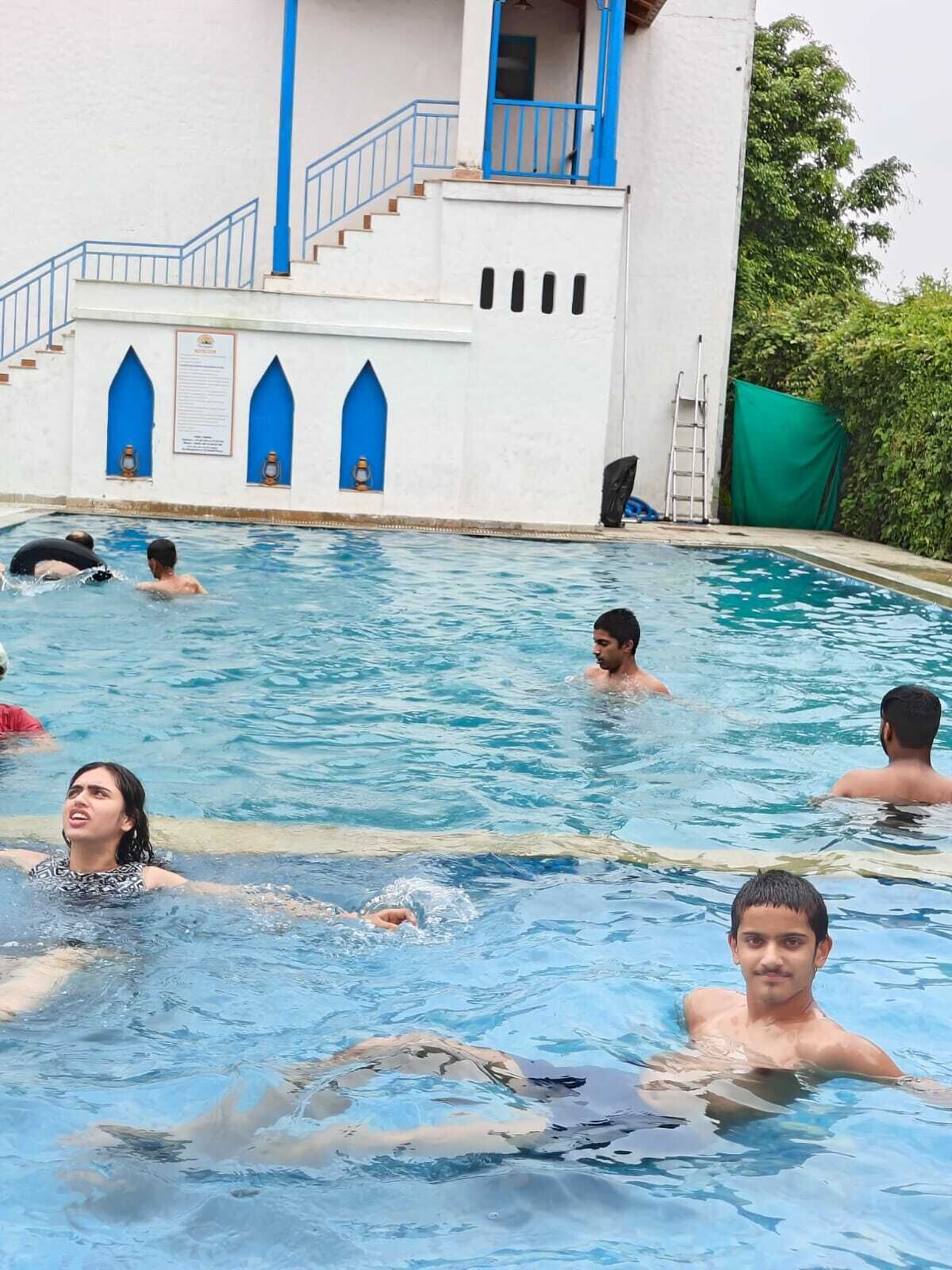 A swimming pool with people in it, a white building with blue accents, and a green netting to the side.