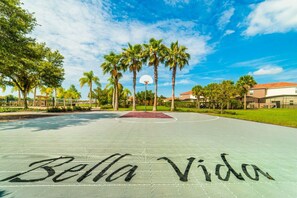 An outdoor basketball court with the words 'Bella Vida' painted on the surface, surrounded by palm trees and buildings. - Aco7947ha - Bella Vida Resort - 4 Bed 3 Baths Townhome (Kissimmee)