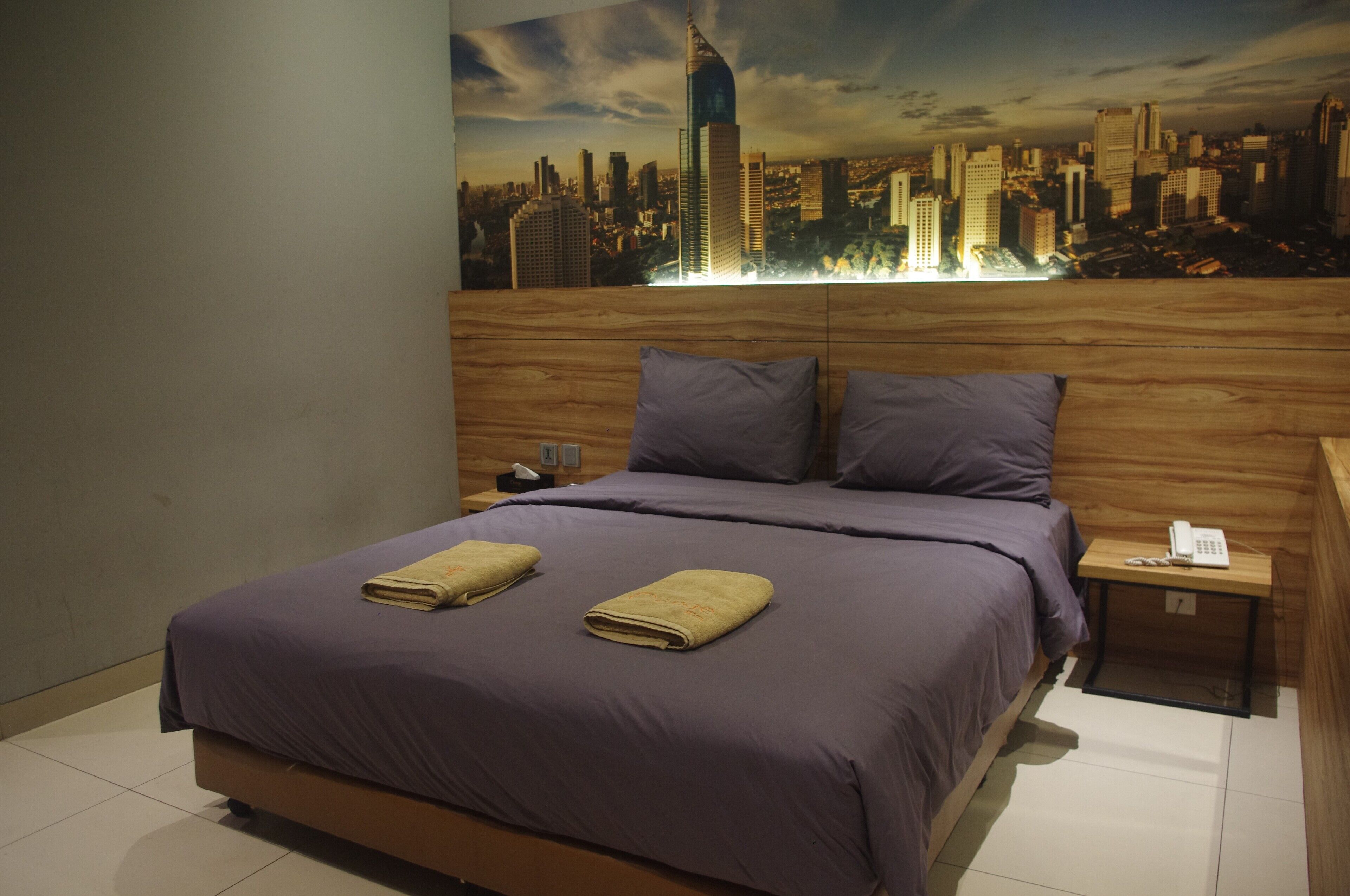 Habitación de hotel con una cama grande, dos almohadas, cabecera de madera, mesita de noche con teléfono y un mural de un paisaje urbano en la pared.
