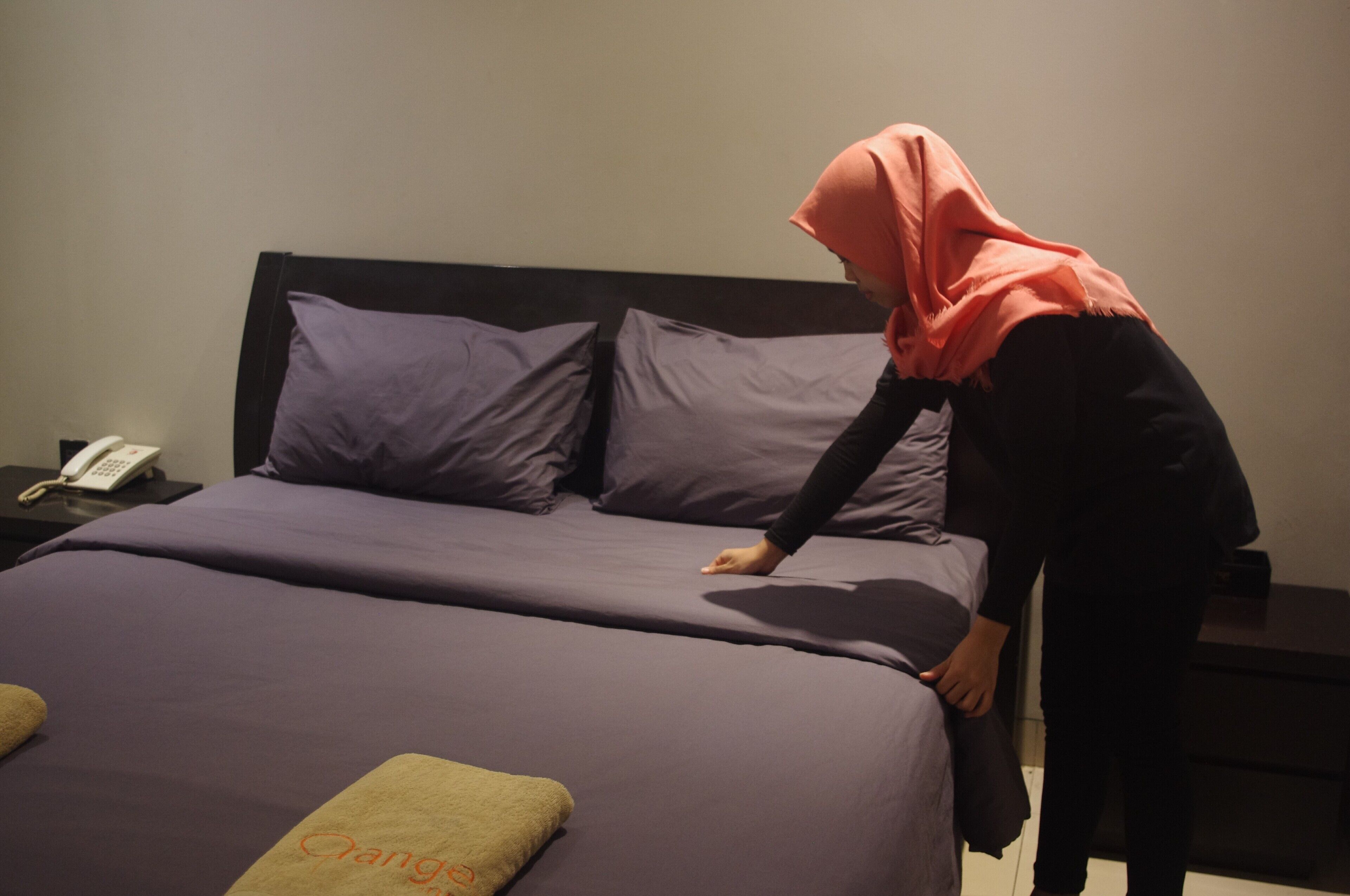 A neatly made bed with purple bedding and a green pillow with the text "Qang" on it.