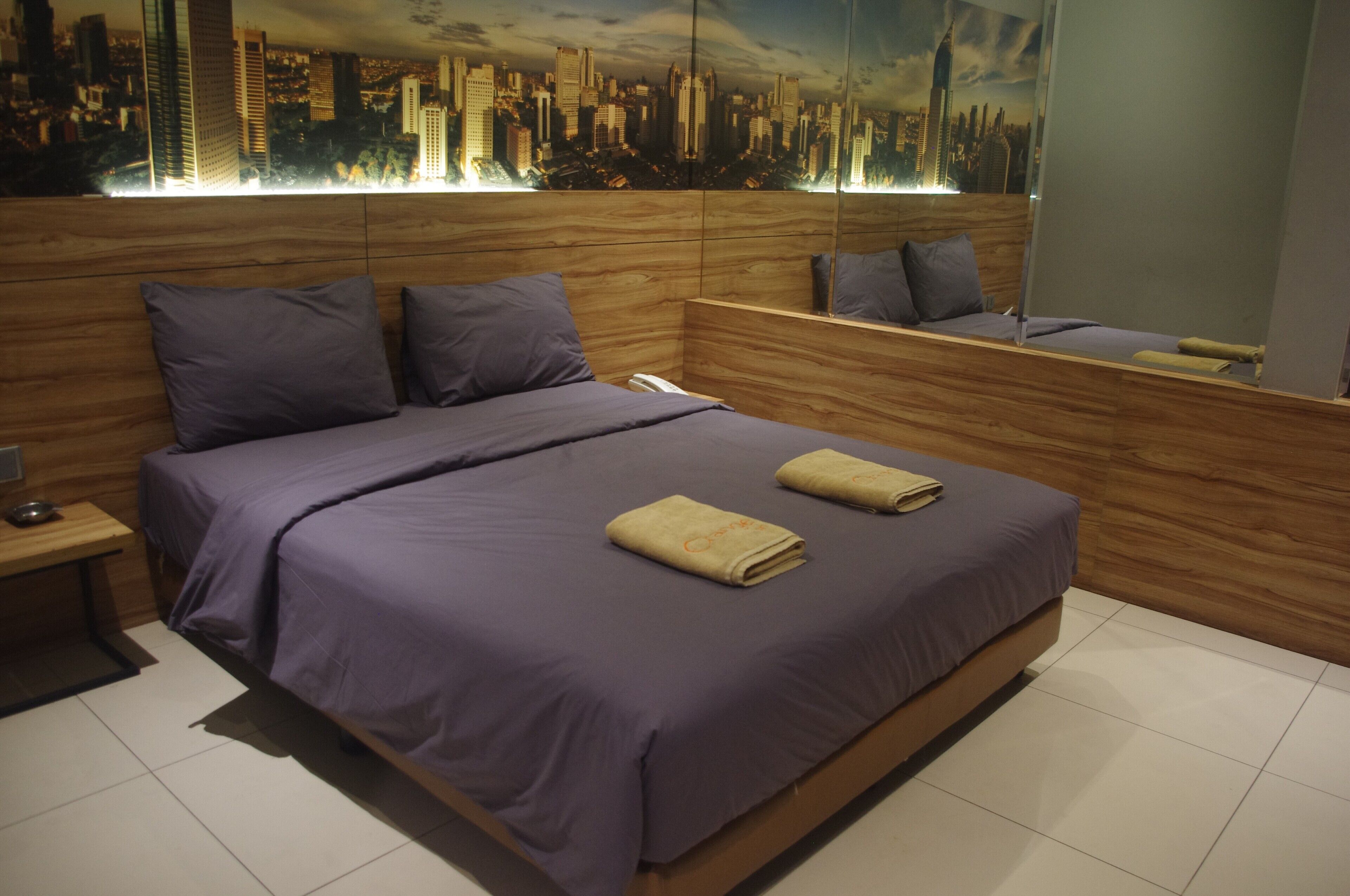 A modern bedroom with a cityscape mural, a wooden headboard, and a bed with purple bedding and towels.