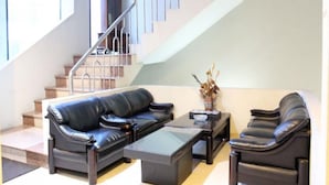 Una sala de estar moderna con un sofá de cuero negro, un sillón a juego y una mesa de centro de vidrio. Se ve una escalera con barandillas de metal al fondo.