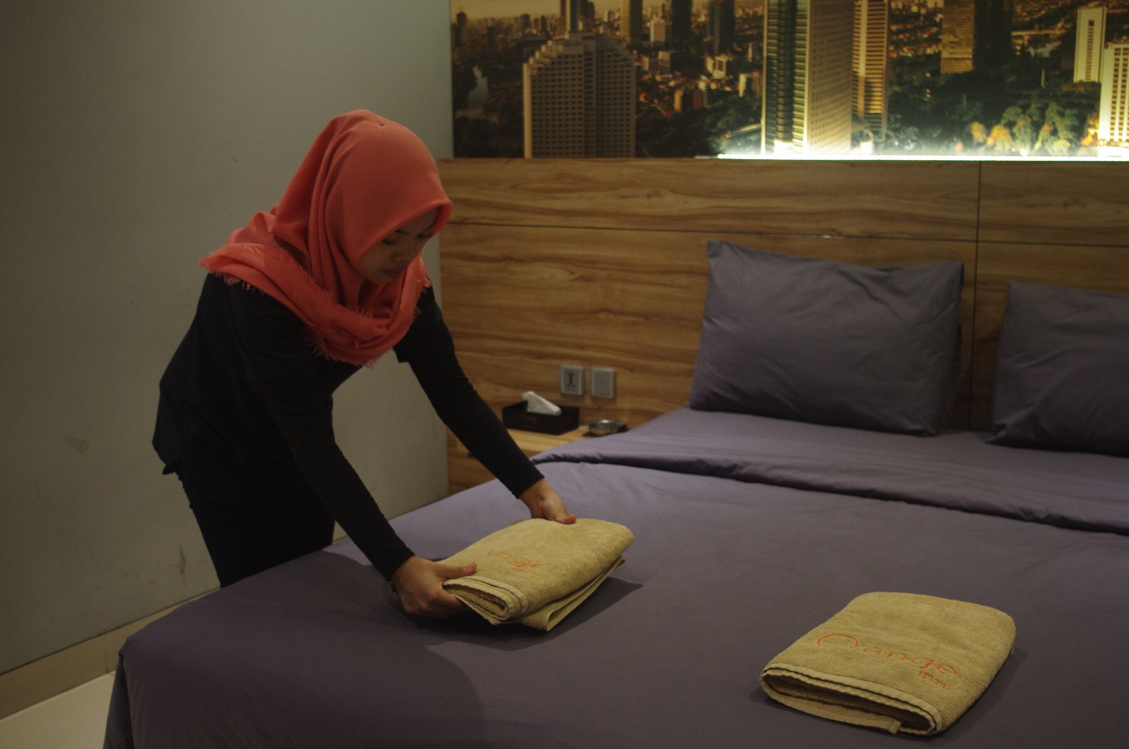 A person folding a towel on a bed with a cityscape view through the window.