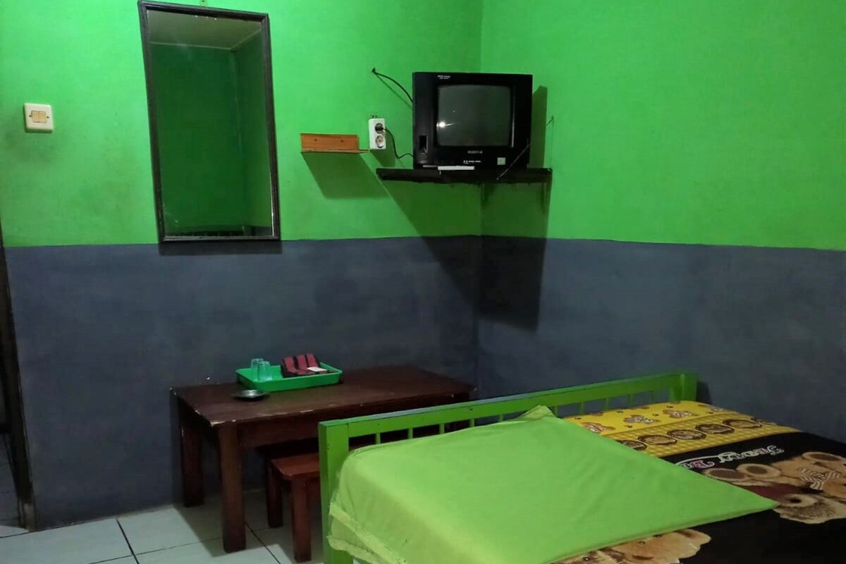 A room with a green and blue wall, a bed with a green bedspread, a small wooden desk with a green tray, a television on a stand, and a mirror on the wall.