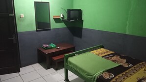 Una habitación pequeña con paredes verdes, una cama individual con manta verde, un escritorio pequeño con silla, un televisor sobre una estantería y una puerta.