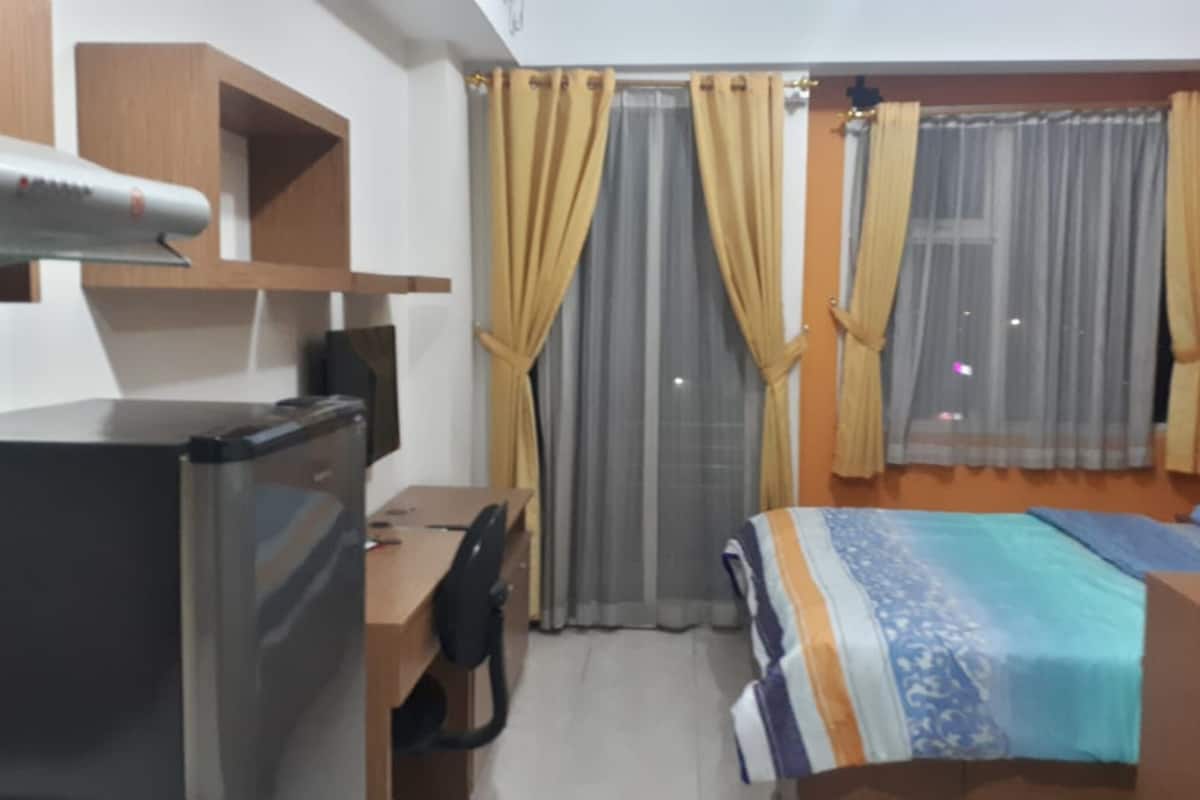 A hotel room with a bed, a desk, a chair, a refrigerator, and curtains.