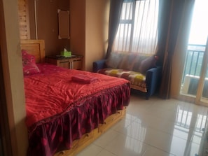 Un dormitorio con cama de madera, una mesita de noche pequeña, una silla y vistas al exterior a través de una ventana con cortinas.