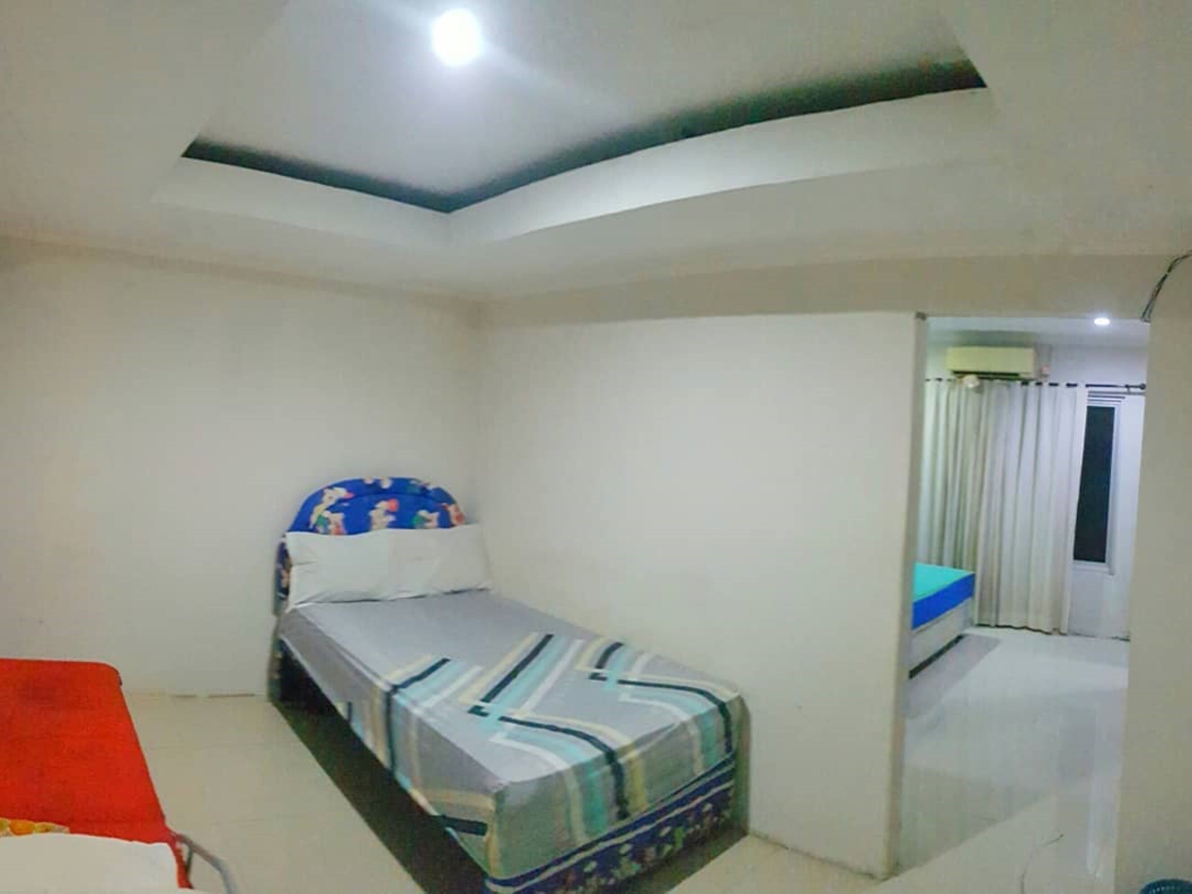 Una habitación pequeña con cama individual, techo y paredes blancos lisos, una silla roja y una ventana con cortinas.