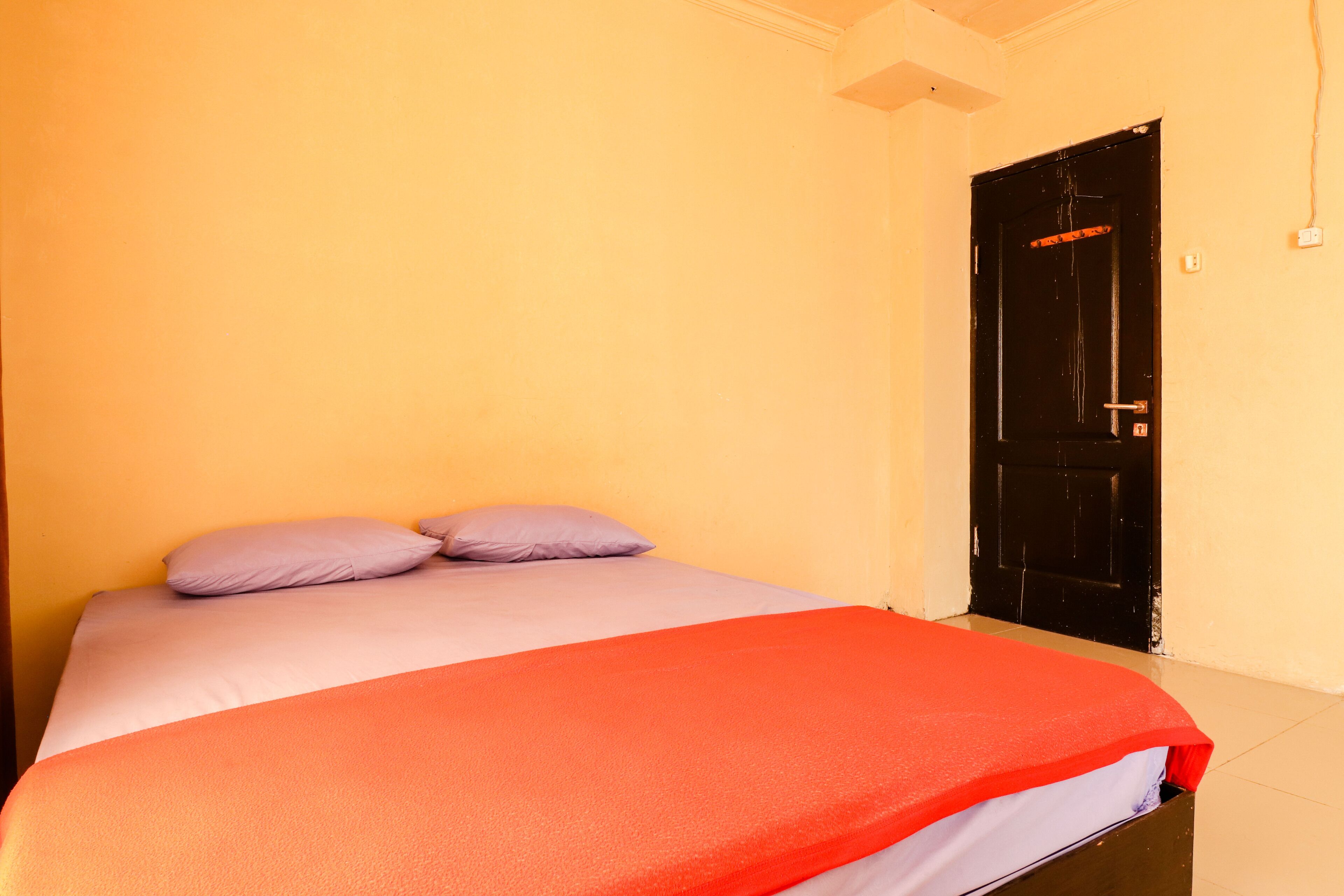 A bed with a red blanket and two pillows, a black door, and a yellow wall.