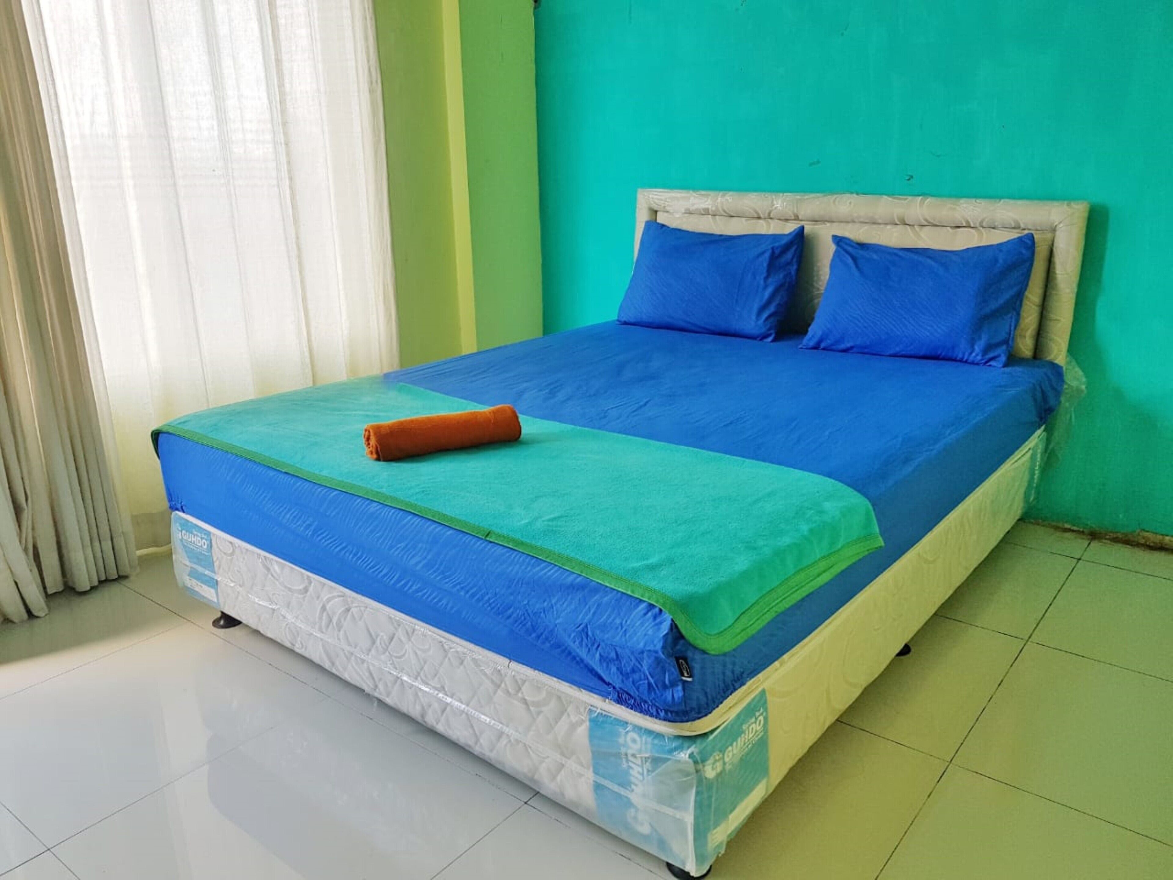 A bed with a blue mattress and green bedspread, a rolled-up orange towel, and a headboard against a teal wall.