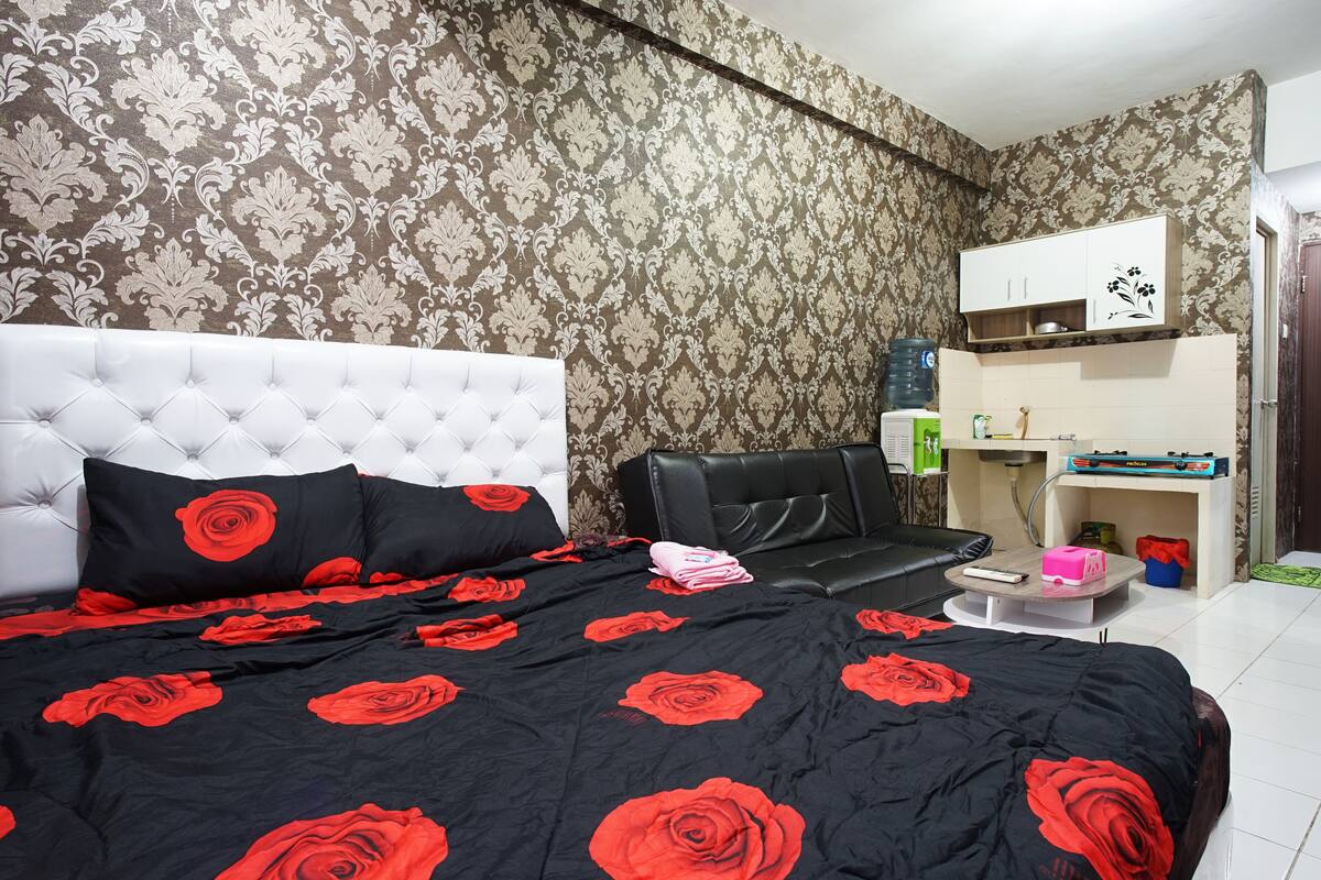 Un dormitorio con cabecera capitonada, una cama con colcha estampada de rosas negras y rojas, un sofá de cuero negro, una mesita con un objeto rosa y un armario blanco con diseño floral.
