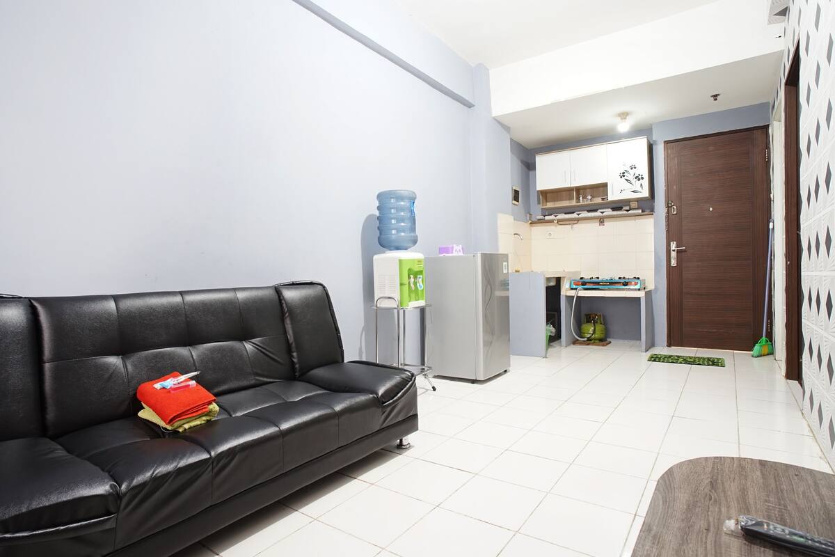 Una sala de estar con un sofá de cuero negro, una mesita de madera, un refrigerador, un dispensador de agua y una zona de cocina con armarios y un microondas.