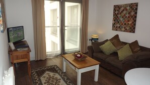 A living room with a television, a wooden side table, a brown sofa with patterned cushions, a small white table, and a patterned rug. - 55 St James Walk (Cheltenham)