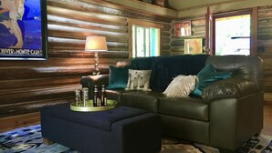 A cozy living room with a dark blue sofa, a matching ottoman, and a patterned rug. A television is mounted on the wall, and a lamp is on a side table. - Stylish Cabin in 12 Acres of Private Forest (Sagle)