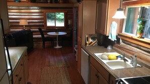 A cozy cabin interior with a dining area, kitchen, and a window overlooking greenery. - Stylish Cabin in 12 Acres of Private Forest (Sagle)