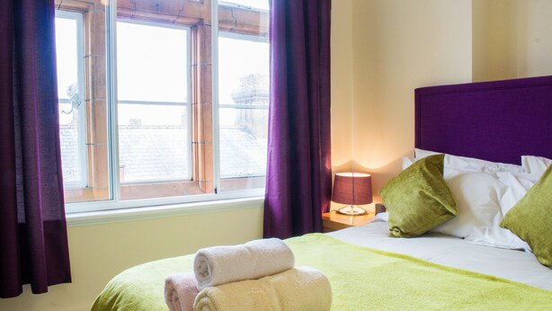 Habitación de hotel con una cama, ropa de cama verde y blanca, una ventana con cortinas moradas y una lámpara en la mesita de noche.