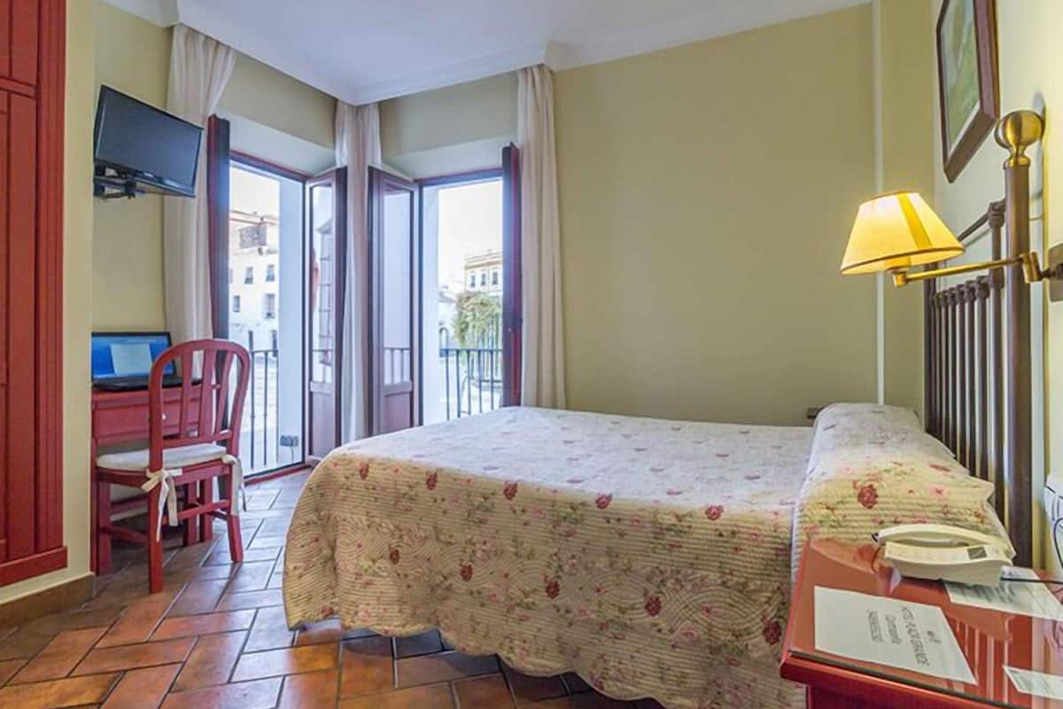 A hotel room with a bed, a desk with a computer, a chair, a lamp, and a view of the outside.