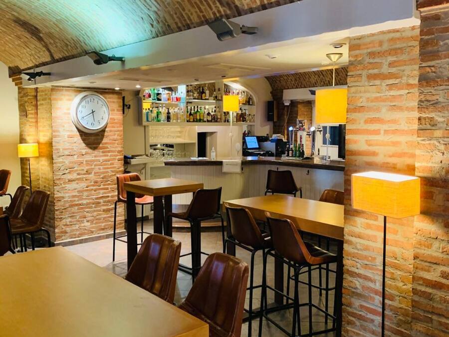 A restaurant with brick walls, wooden tables, and chairs. There is a clock on the wall and a bar area with bottles.
