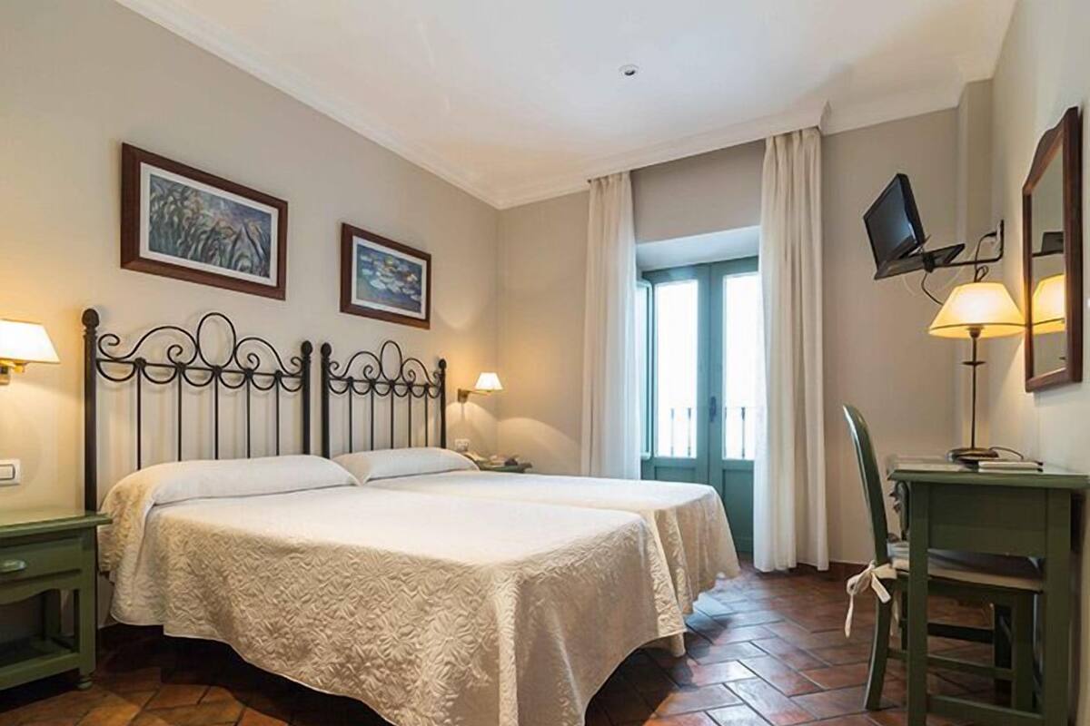 A hotel room with a double bed, two bedside tables with lamps, a desk with a chair, a TV mounted on the wall, and a window with curtains.