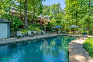 Ein Swimmingpool mit Liegestühlen und Sonnenschirmen, umgeben von Bäumen und einer Holzterrasse.