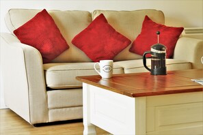 Un canapé beige avec des coussins rouges, une tasse blanche portant l’inscription « LATTE » et une machine à café noire sur une table en bois.