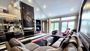 Un salon moderne avec un grand canapé d’angle, une table basse en verre et un téléviseur à écran plat. On y trouve également un coin bar avec des étagères en verre et un fauteuil rouge.