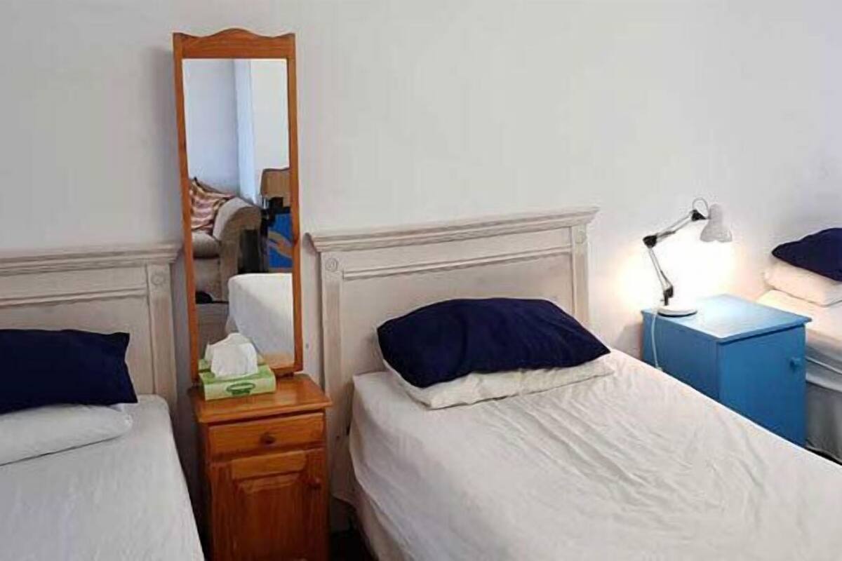Habitació d'hotel amb quatre llits individuals, cadascun amb matalàs blanc i almoina blava fosca. Hi ha una nituixi de fusta amb una làmpada a un costat i un petit armari blau a l'altre. Un mirall està fixat a la paret sobre el llit més a l'esquerra.