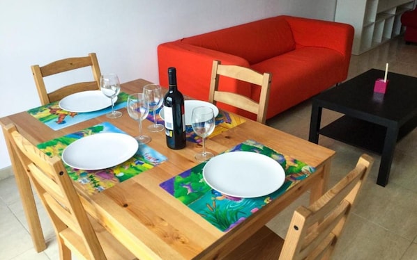 A wooden dining table set with plates, wine glasses, and a bottle, with a red sofa and a black table in the background.